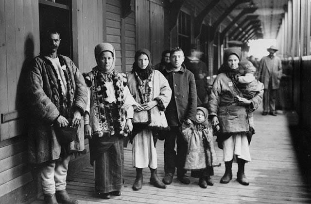 Galician immigrants, including women in headscarves and a small child, standing on a covered wooden platform beside a railway car, about 1911.
