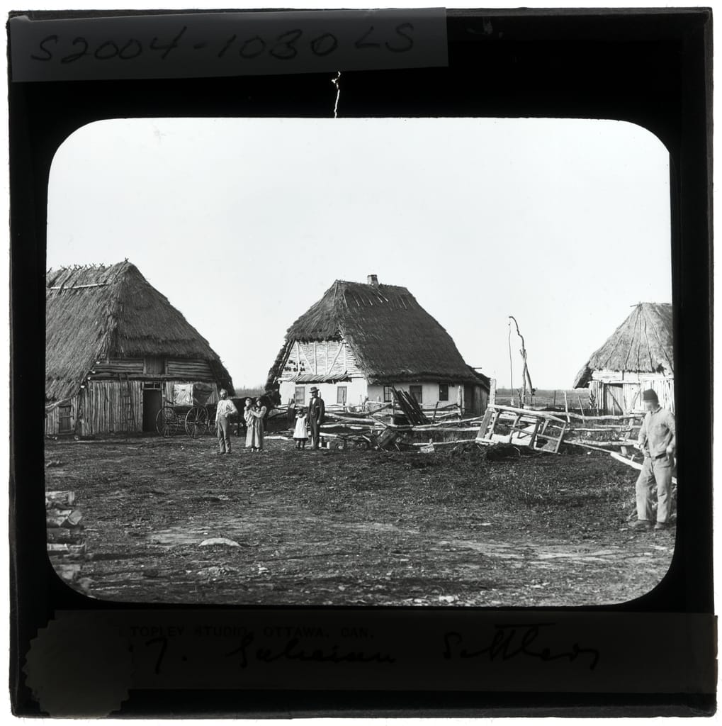 Three thatched log houses on open ground, with a family standing in the yard. A lantern slide labelled Galician Settlers.