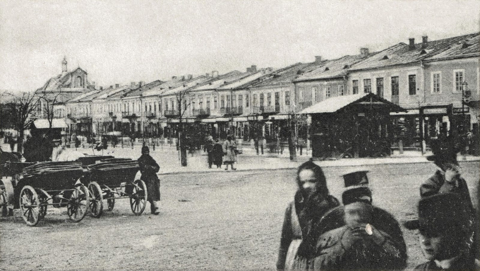 A wide unpaved market square lined with two-storey buildings, carts, and people in heavy coats, in Kolomyia between 1900 and 1904.