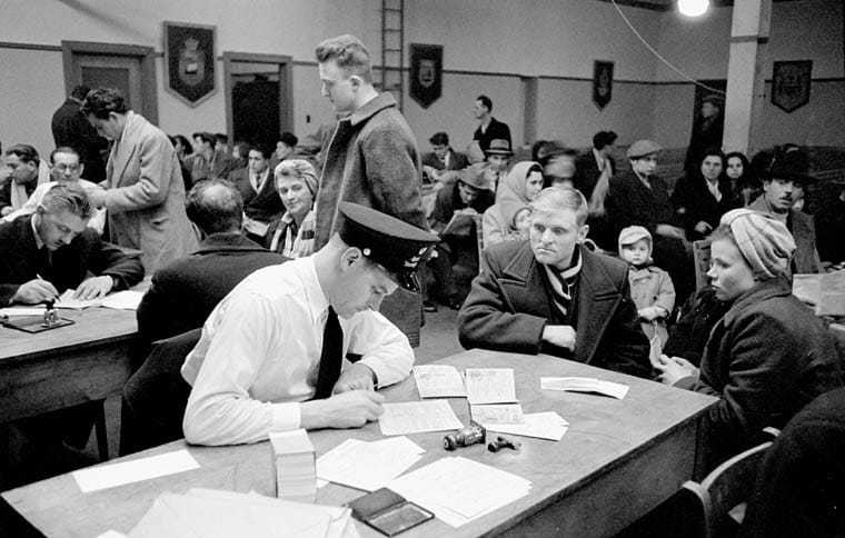 Immigrants seated at long tables in a large hall while an officer in a peaked cap checks papers, Pier 21, Halifax, May 1952.