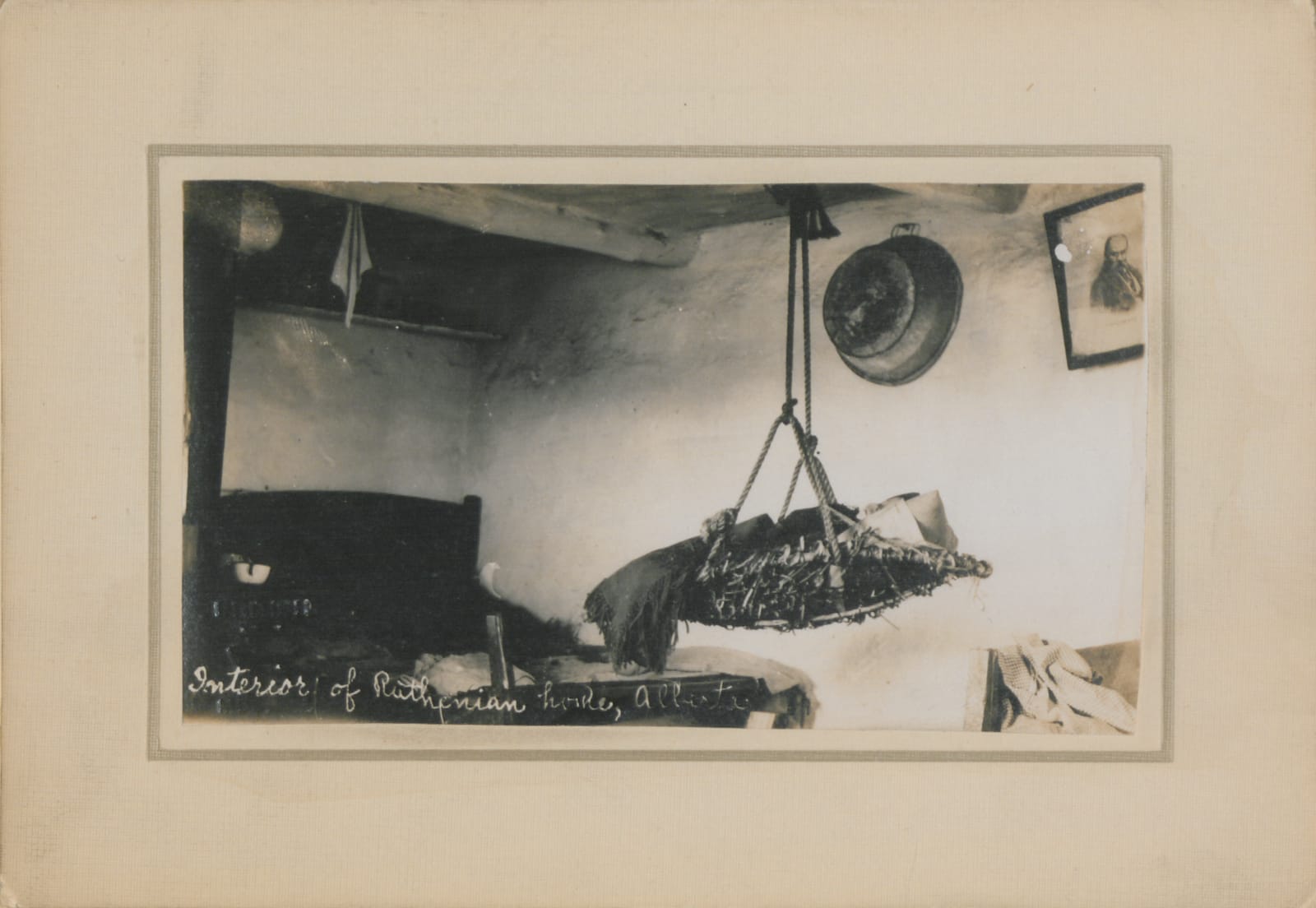 A dim room with clay or plaster walls, a hanging cradle, a basin, and a dark cupboard. Handwritten caption: Interior of Ruthenian home, Alberta.