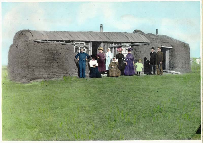A long low sod house on the prairie with a group of people in early twentieth-century dress standing at the doors.