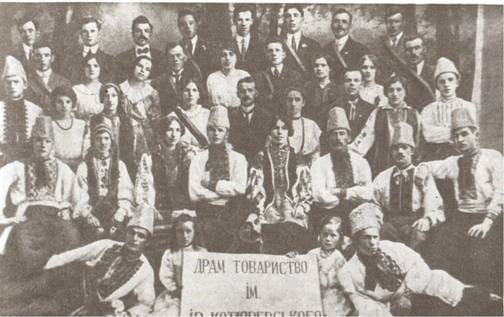 A large group in embroidered shirts and costumes posed in rows, with a placard in Ukrainian for a dramatic society, 1914.
