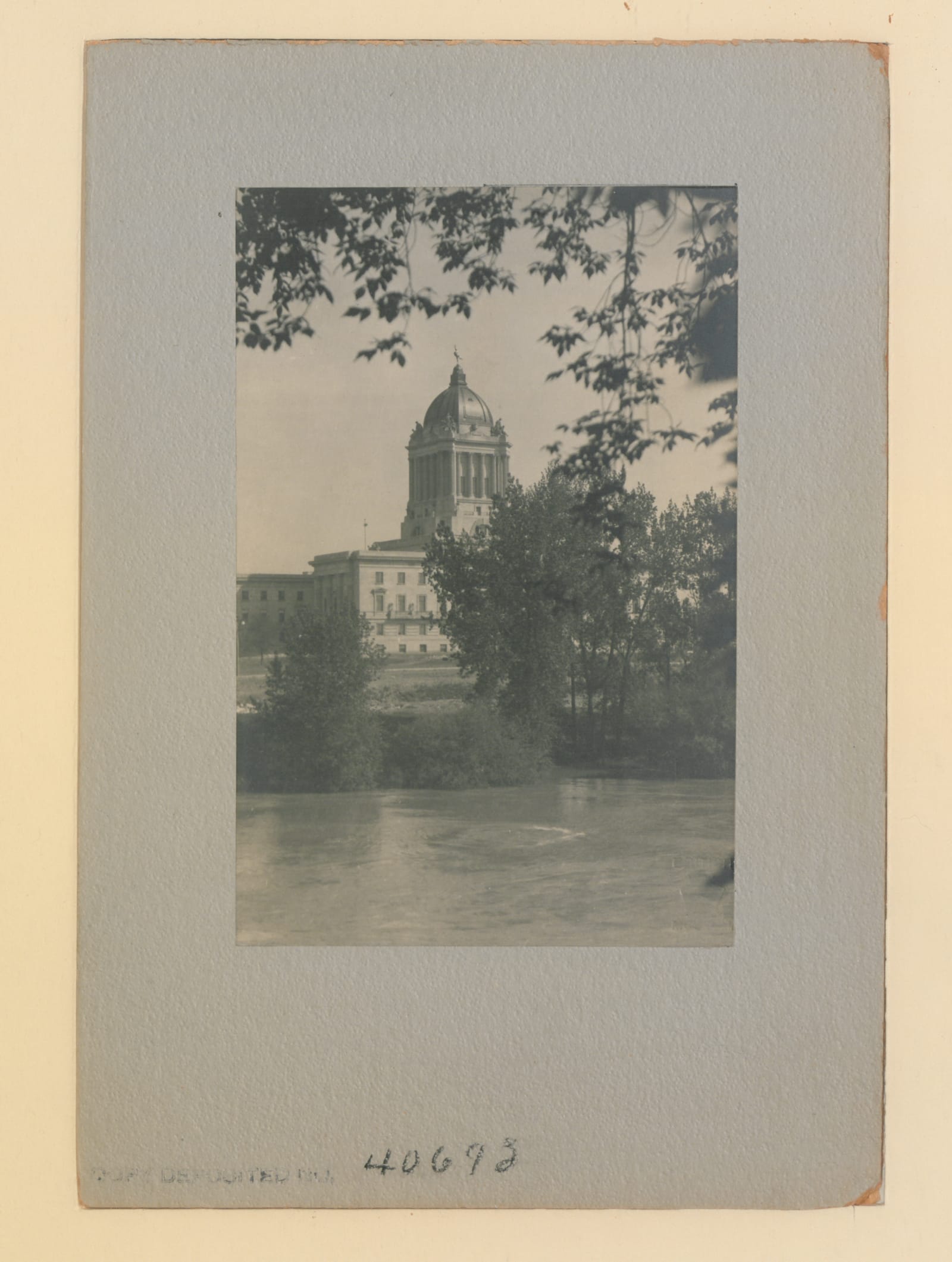 The domed Manitoba Legislative Building seen across the river, framed by trees, in a mounted 1922 photograph.